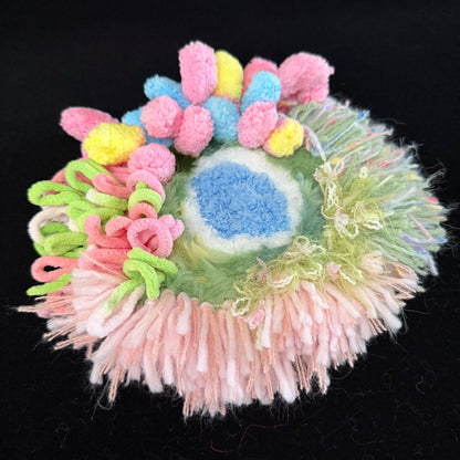 Handmade Blossom Puddle pastel pom-pom crochet coaster angled side view showing layered pink blue yellow pom-poms green curly yarn and rainbow fringe edge on black background – Sylva Garden