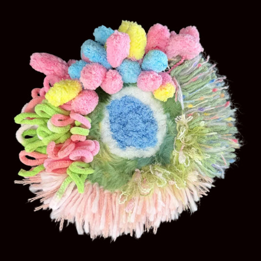 Handmade Blossom Puddle pastel pom-pom crochet coaster overhead view showing blue center with pink blue yellow pom-poms green curly yarn accents and rainbow fringe border on black background – Sylva Garden