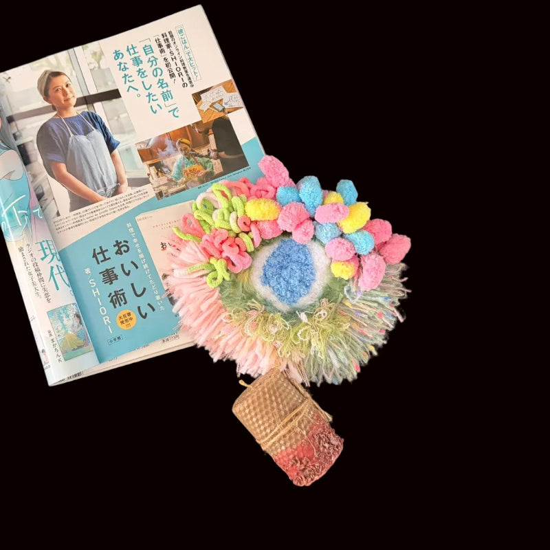 Handmade Blossom Puddle pastel pom-pom crochet coaster styled with Japanese magazine and beeswax candle on black background, lifestyle view – Sylva Garden