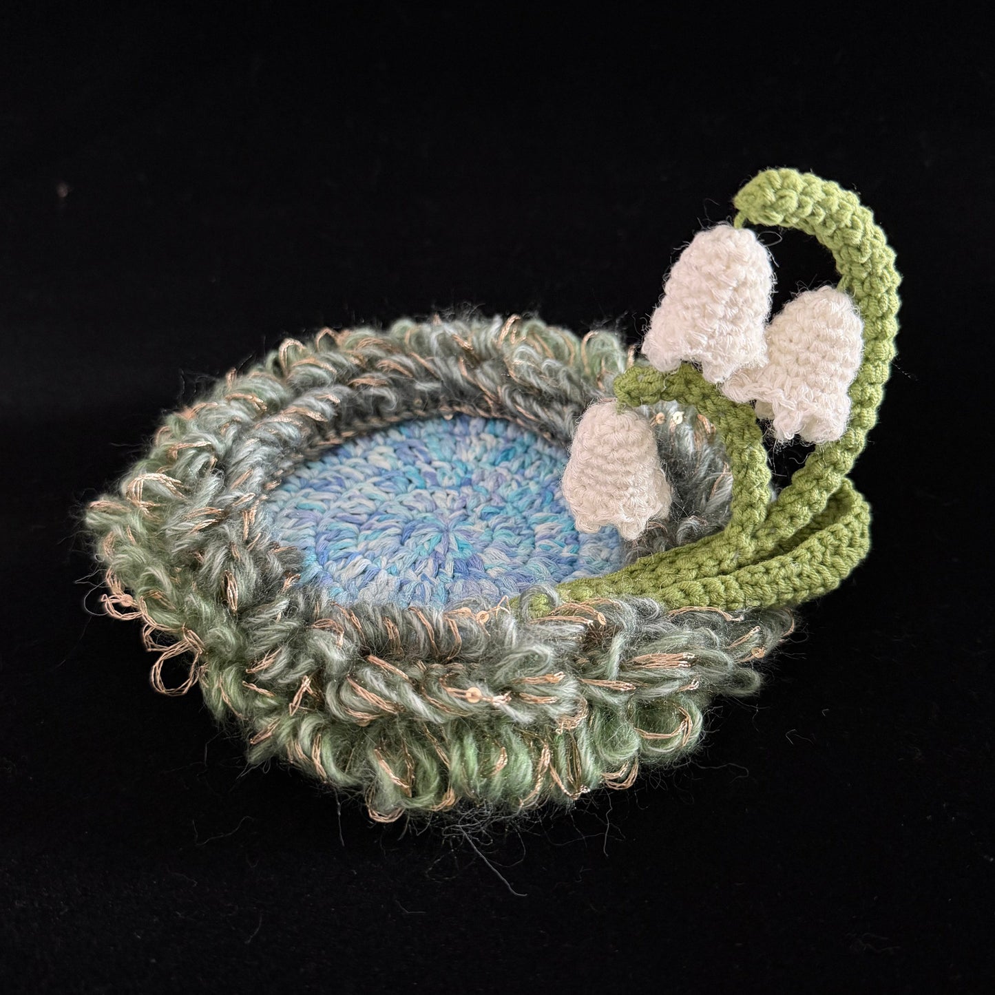 Handmade Dewbell lily of the valley crochet moss coaster angled overhead view showing blue center green moss border and white bell blooms on black background – Sylva Garden