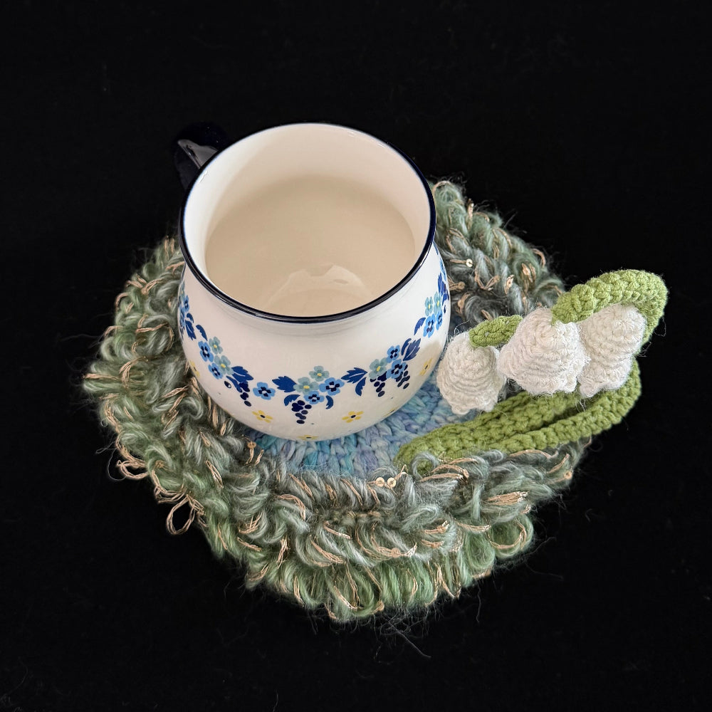 Handmade Dewbell lily of the valley crochet moss coaster with floral enamel mug overhead view showing green moss texture and white bell bloom accent on black background – Sylva Garden
