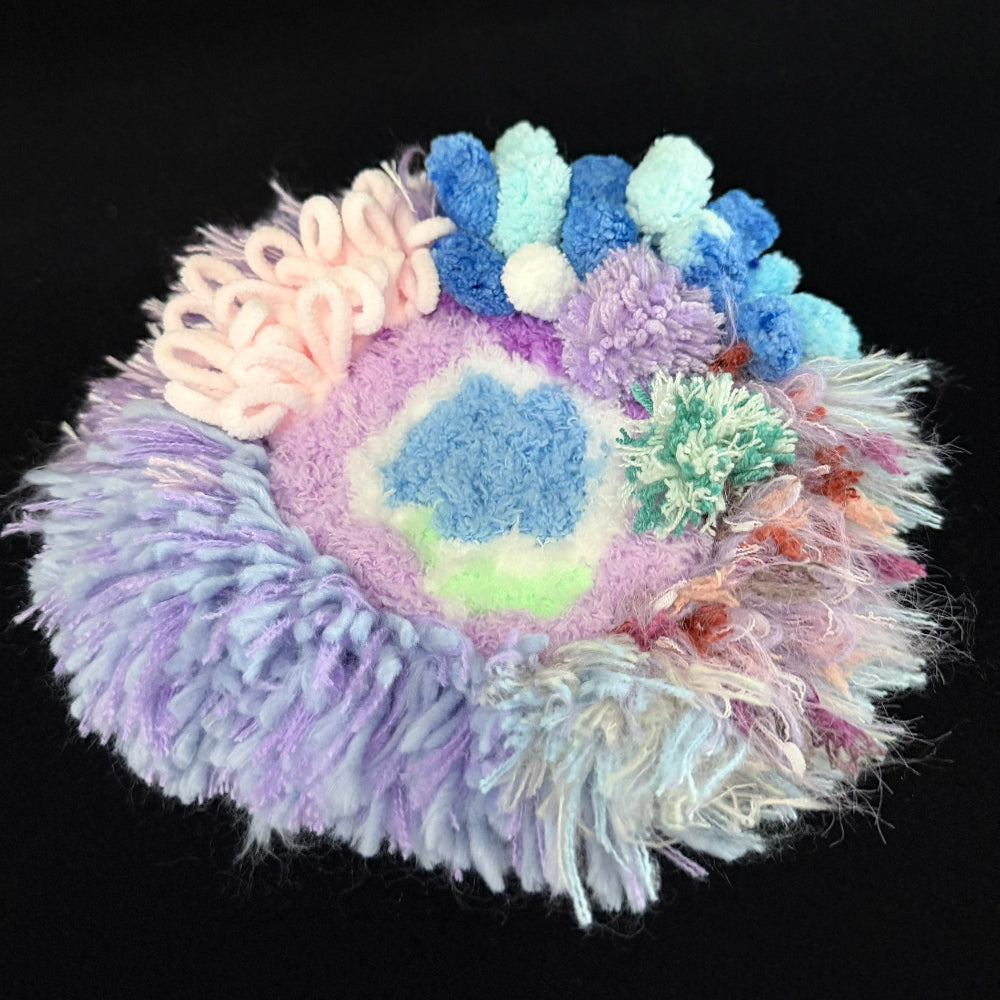 Handmade Dreamy Bog lavender blue pink coral pom-pom crochet coaster angled view showing blue center lavender pink pom-pom clusters and multicolor fringe border on black background – Sylva Garden