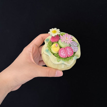 Handmade clay succulent arrangement in green lotus-shaped pot held in hand showing side profile with layered pink lithops yellow succulent white water lily and pink daisy – Sylva Garden