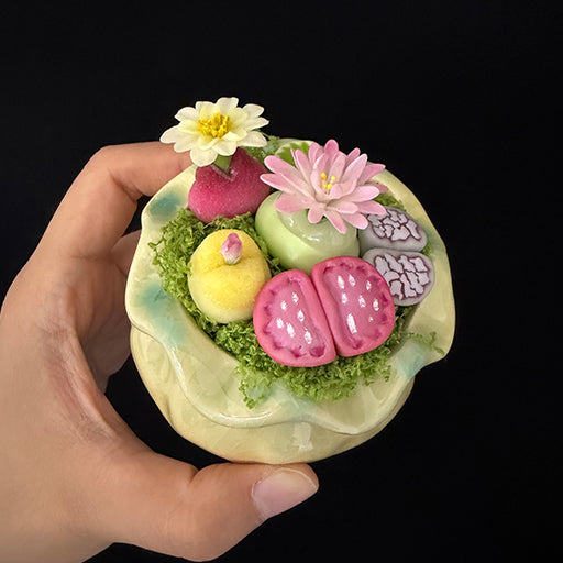 Handmade clay succulent arrangement in green lotus-shaped pot held in hand angled top view showing full pink lithops yellow succulent pink daisy white water lily green cactus and moss detail – Sylva Garden