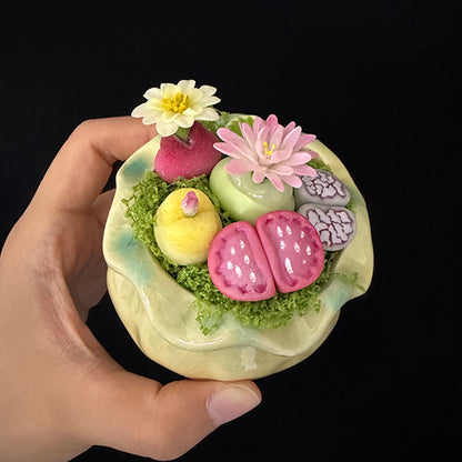 Handmade clay succulent arrangement in green lotus-shaped pot held in hand angled top view showing full pink lithops yellow succulent pink daisy white water lily green cactus and moss detail – Sylva Garden