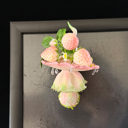 Handmade milky spore strawberry mushroom resin clay magnet with pink ruffled cap and strawberry accents attached to grey metal frame, lifestyle view – Sylva Garden