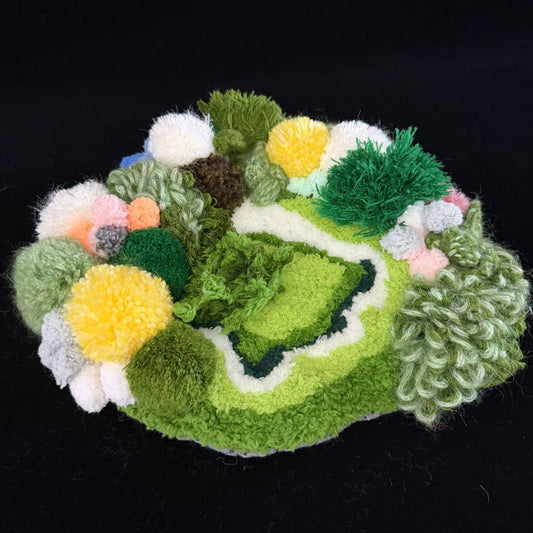 Handmade Moss Wanderer green multi-texture pom-pom crochet coaster angled side view showing layered dark green light green yellow white and brown pom-pom clusters with curly yarn border on black background – Sylva Garden