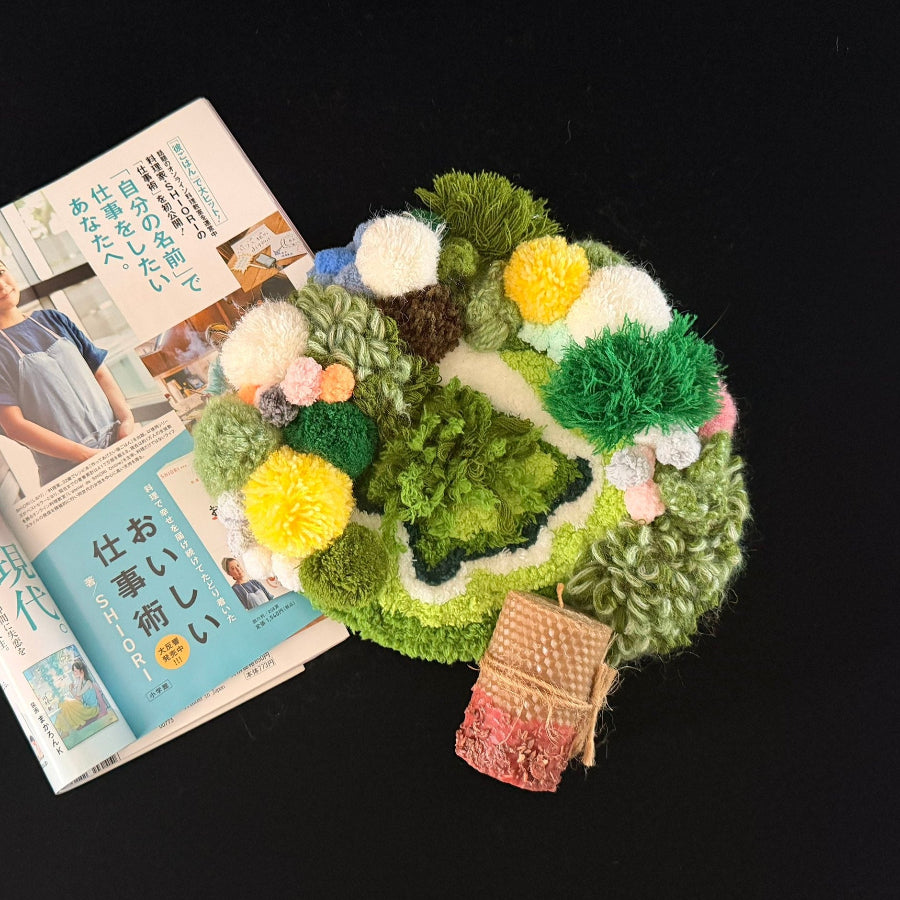 Handmade Moss Wanderer green multi-texture pom-pom crochet coaster styled with Japanese magazine and beeswax candle on black background, overhead lifestyle view – Sylva Garden