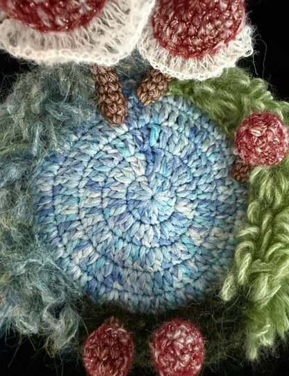Handmade Pixie Ring red lace mushroom crochet moss coaster close-up showing blue spiral crochet center texture with red mushroom stems and green moss border on black background – Sylva Garden