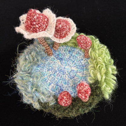 Handmade Pixie Ring red lace mushroom crochet moss coaster overhead view showing blue center with red lace mushroom cluster and curly green moss border on black background – Sylva Garden