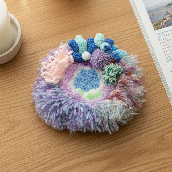 Handmade Dreamy Bog lavender blue pink coral pom-pom crochet coaster with fluffy fringe border, overhead lifestyle view on wooden table with white cup and Japanese magazine – Sylva Garden