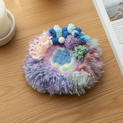 Handmade Dreamy Bog lavender blue pink coral pom-pom crochet coaster with fluffy fringe border, overhead lifestyle view on wooden table with white cup and Japanese magazine – Sylva Garden