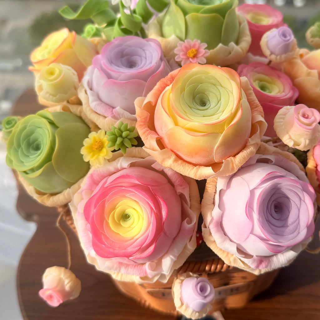 Rose Succulent Carnival handmade clay ranunculus and rose succulent arrangement angled view showing full pastel bouquet with green foliage and daisy accents in woven basket on wooden desk – Sylva Garden