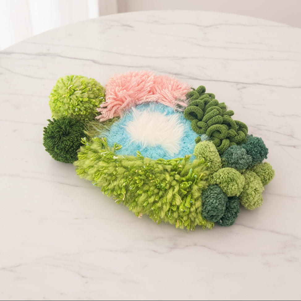 Summer River handmade crochet moss coaster side view on marble surface showing green textured base with blue water center pink arch and mixed green pompom accents – Sylva Garden