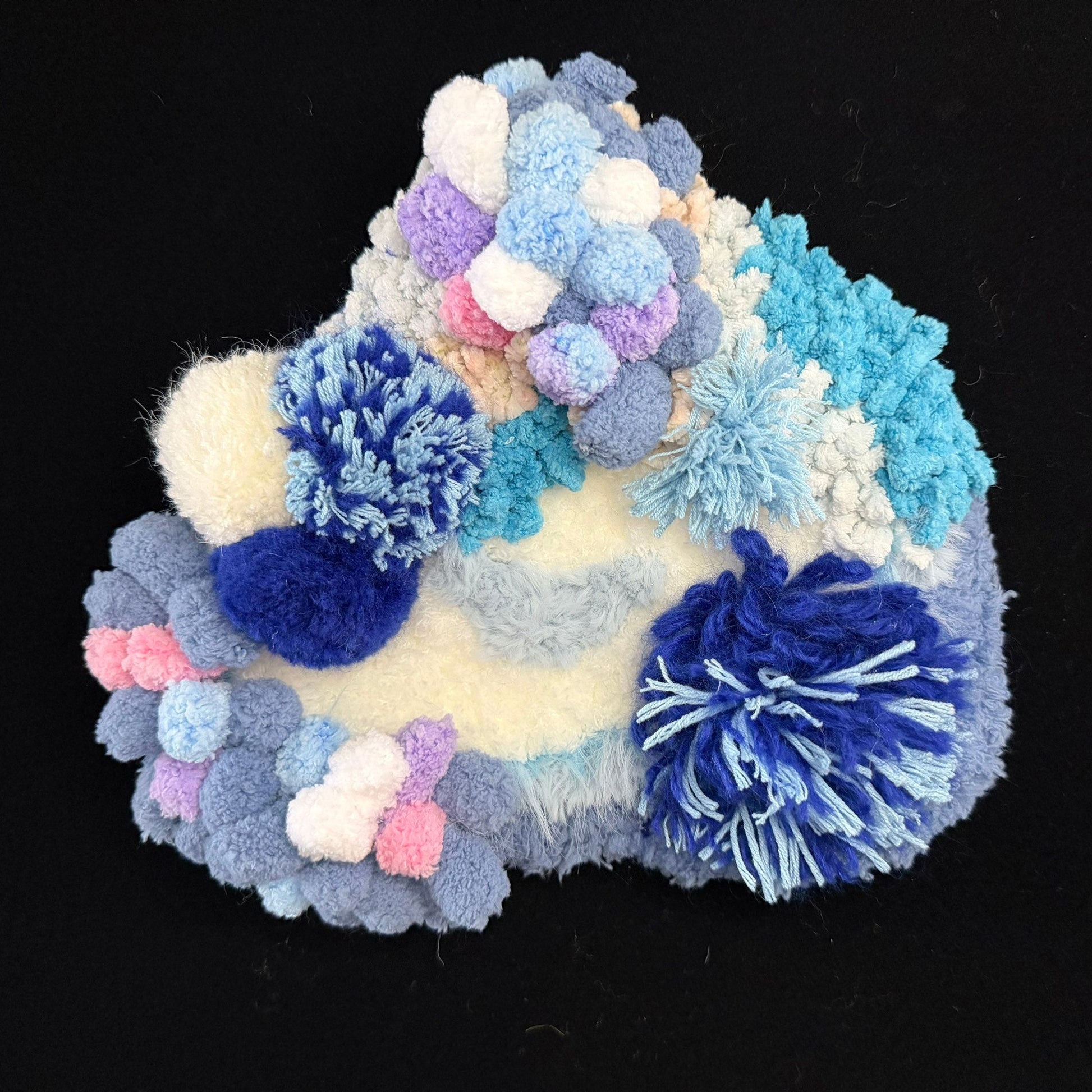 Handmade Tidal Moss blue white gradient pom-pom crochet coaster showing navy teal lavender pink and cream pom-pom clusters, angled view on black background – Sylva Garden
