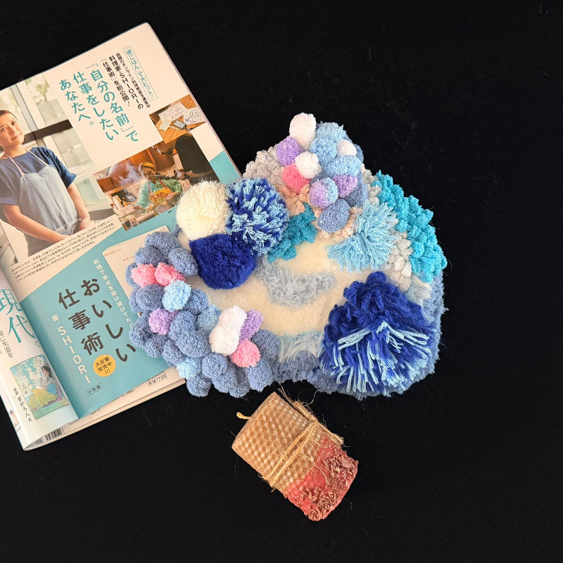 Handmade Tidal Moss blue white gradient pom-pom crochet coaster styled with Japanese magazine and beeswax candle on black background, lifestyle view – Sylva Garden