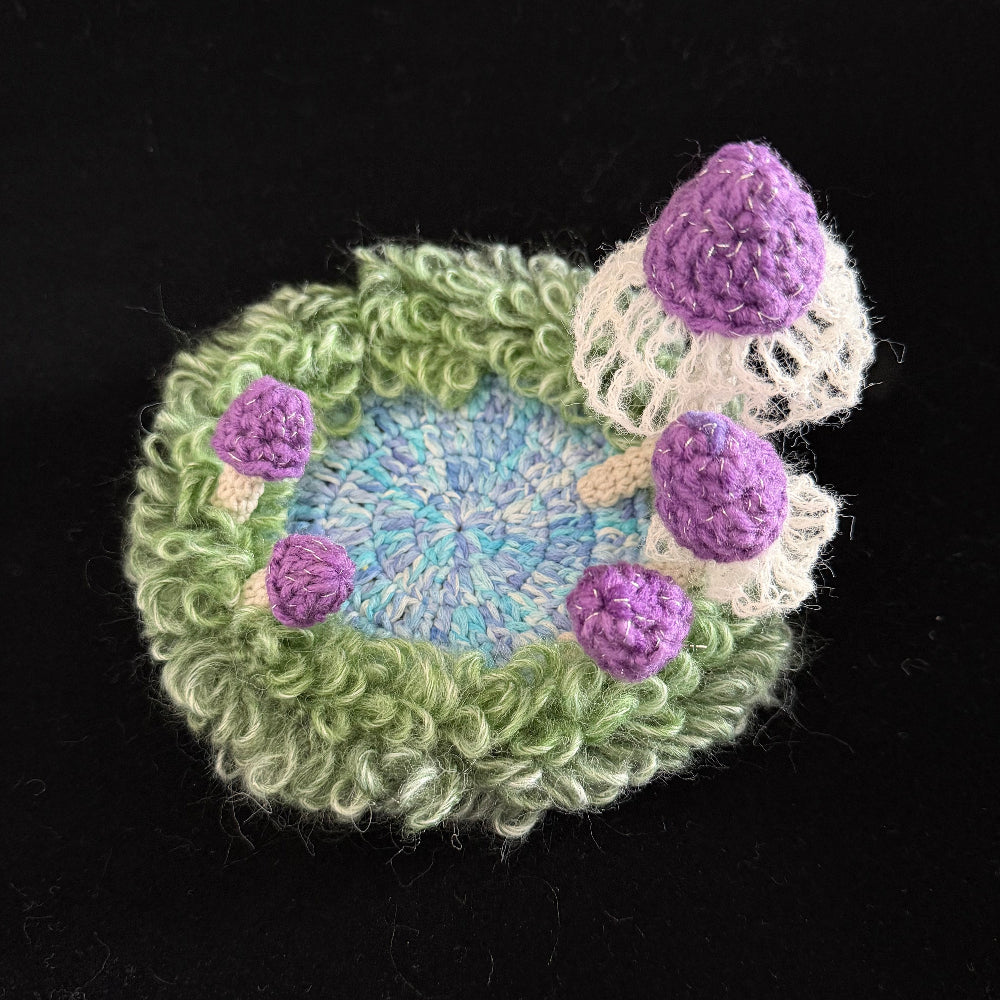 Handmade Violet Veil purple mushroom crochet moss coaster overhead view showing blue center green moss ring and purple mushroom caps with white lace veils on black background – Sylva Garden