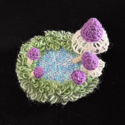 Handmade Violet Veil purple mushroom crochet moss coaster overhead view showing blue center green moss ring and purple mushroom caps with white lace veils on black background – Sylva Garden
