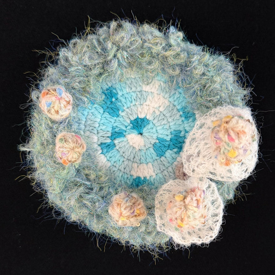 Handmade Whisper Cap ivory lace mushroom crochet moss coaster overhead view showing teal blue and white center with ivory lace mushroom cluster and fluffy moss border on black background – Sylva Garden
