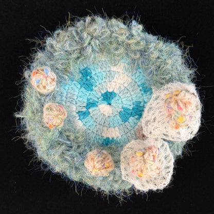 Handmade Whisper Cap ivory lace mushroom crochet moss coaster overhead view showing teal blue and white center with ivory lace mushroom cluster and fluffy moss border on black background – Sylva Garden